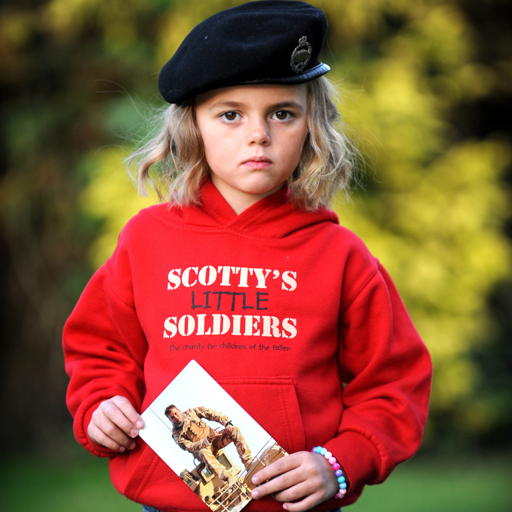 Get support for bereaved military children - Scotty's Little Soldiers support children bereaved of a parent who served in the British Armed Forces. Scotty Member in red hoodie holds a photo of her father, Corporal Lee Scott