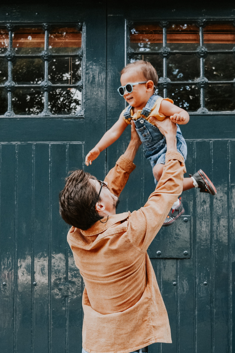 Father in orange shirt lifts child in dungarees and sunglasses into the air in front of green wooden door
