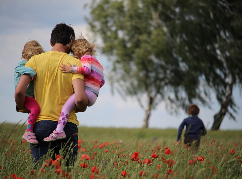 Father wearing yellow shirt walking through field of poppies towards the trees while holding two children wearing rainbow outfits, another child runs off in the distance