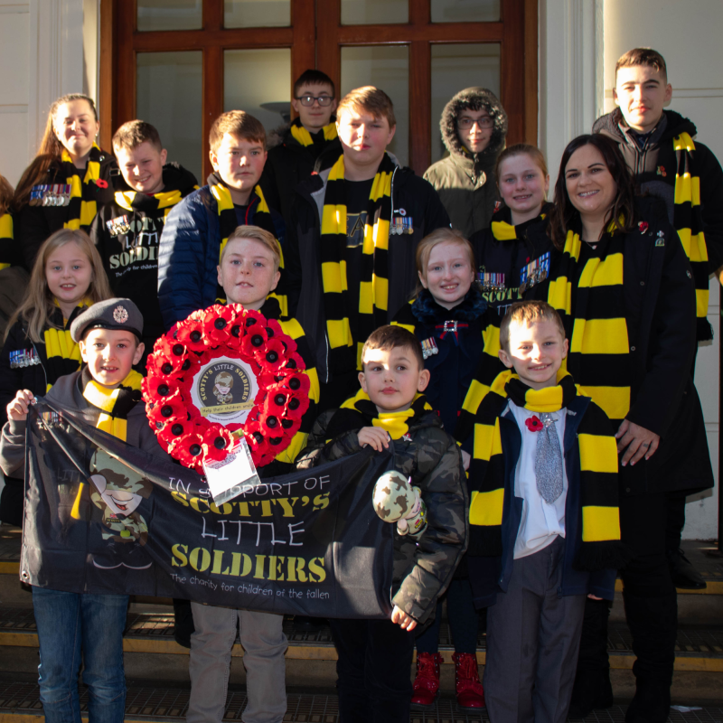 Charity founder Nikki Scott with Scotty Members before Remembrance parade holding up poppy wreath and Scotty's flag, wearing yellow and black scarves
